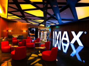 Modern cinema lobby featuring a glowing IMAX sign, geometric ceiling lights, and red lounge chairs, KhaleejFusion.