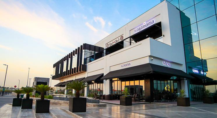 Modern outdoor commercial plaza featuring Godiva and Wagamama storefronts at dusk, KhaleejFusion